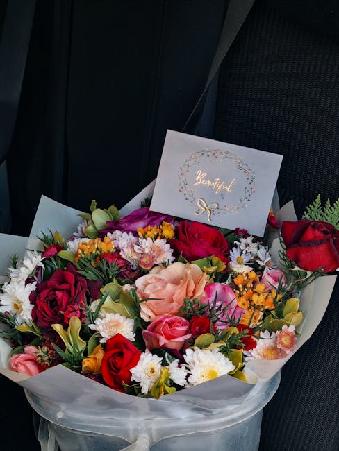 A vibrant floral bouquet featuring red roses, peach and pink carnations, white daisies, yellow daisies, and small purple and yellow filler flowers, arranged with green foliage and wrapped in transluce