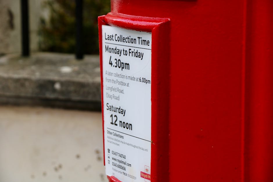 A close-up view of a red postbox with a white schedule notice attached, displaying collection times from Monday to Friday at 4:30 pm and on Saturday at noon. The background includes a blurred stone st