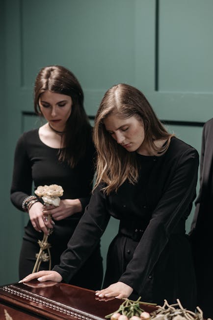 Two women dressed in black mourning attire are gently touching a polished wooden casket during a funeral service. One woman holds a small floral arrangement featuring white roses and greenery, while t